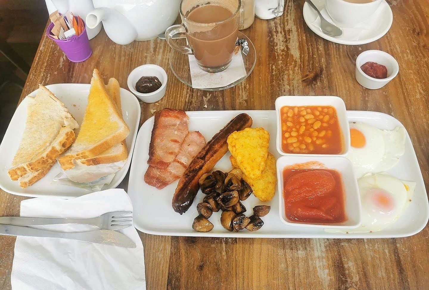 A cooked breakfast on a white tray: bacon, sausage, a hash brown, mushrooms, beans, tomatoes and two fried eggs, with toast and a glass of tea on a wooden table