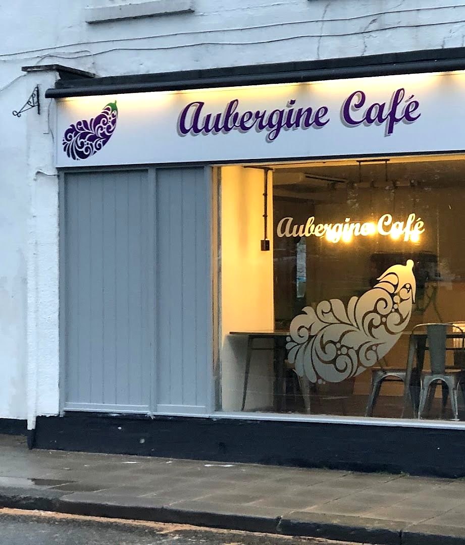 The front of Aubergine Café: a white sign with a purple aubergine and the name in purple script, grey panels, and a window with the name lit up inside
